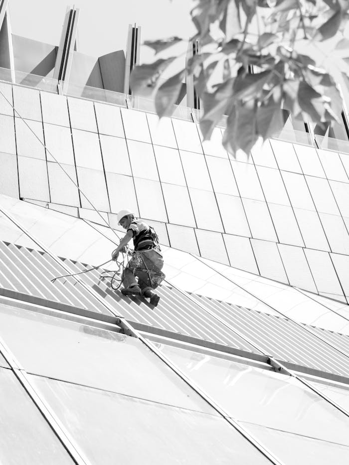 A worker cleans the glass facade of a modern building using ropes for safety, on a bright day.