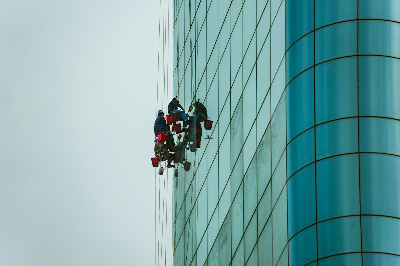 Window cleaners suspended on ropes working on a tall glass building facade.